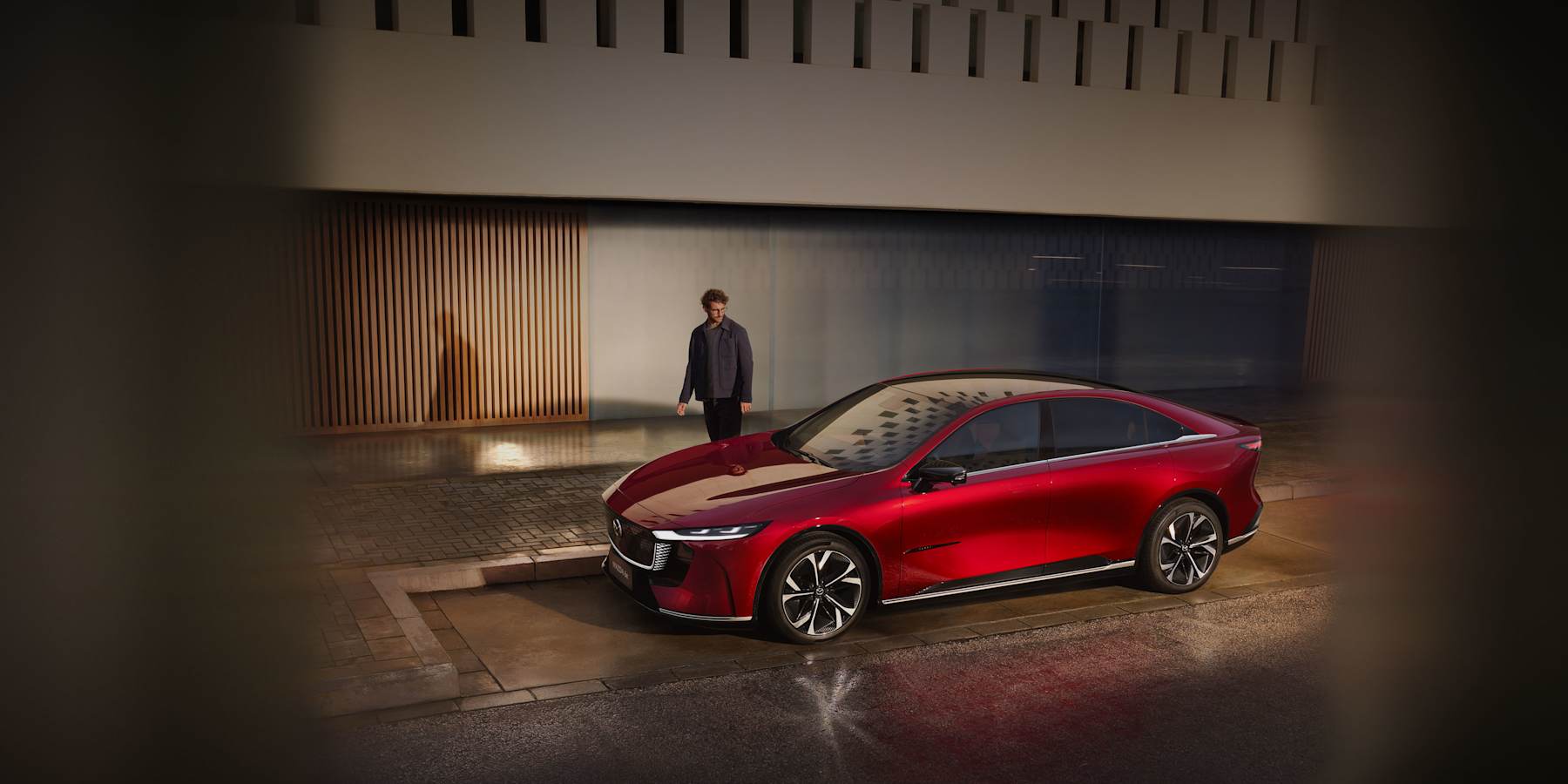 En röd Mazda 6e ståendes på en asfalterad parkering. En man står bredvid bilen och tittar på den.